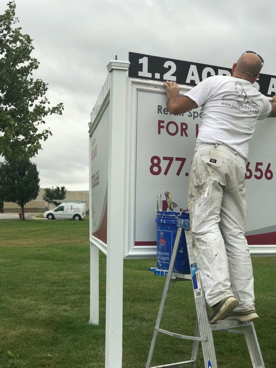 Man on a ladder painting a signboard outdoors beside a white sign with red and black text