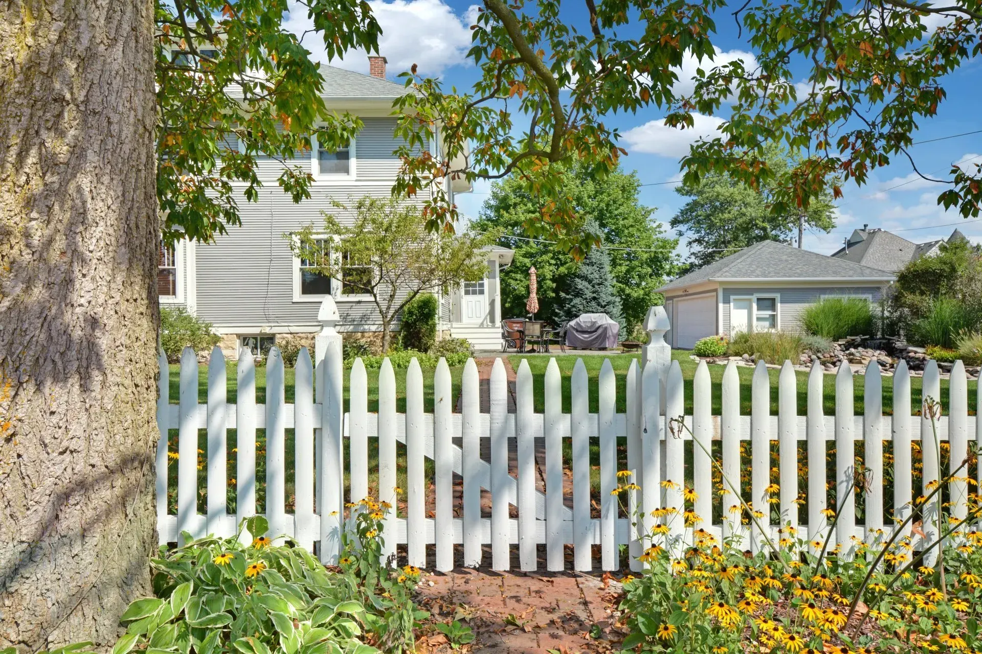 White picket fence in front of a gray house and garden under a leafy tree