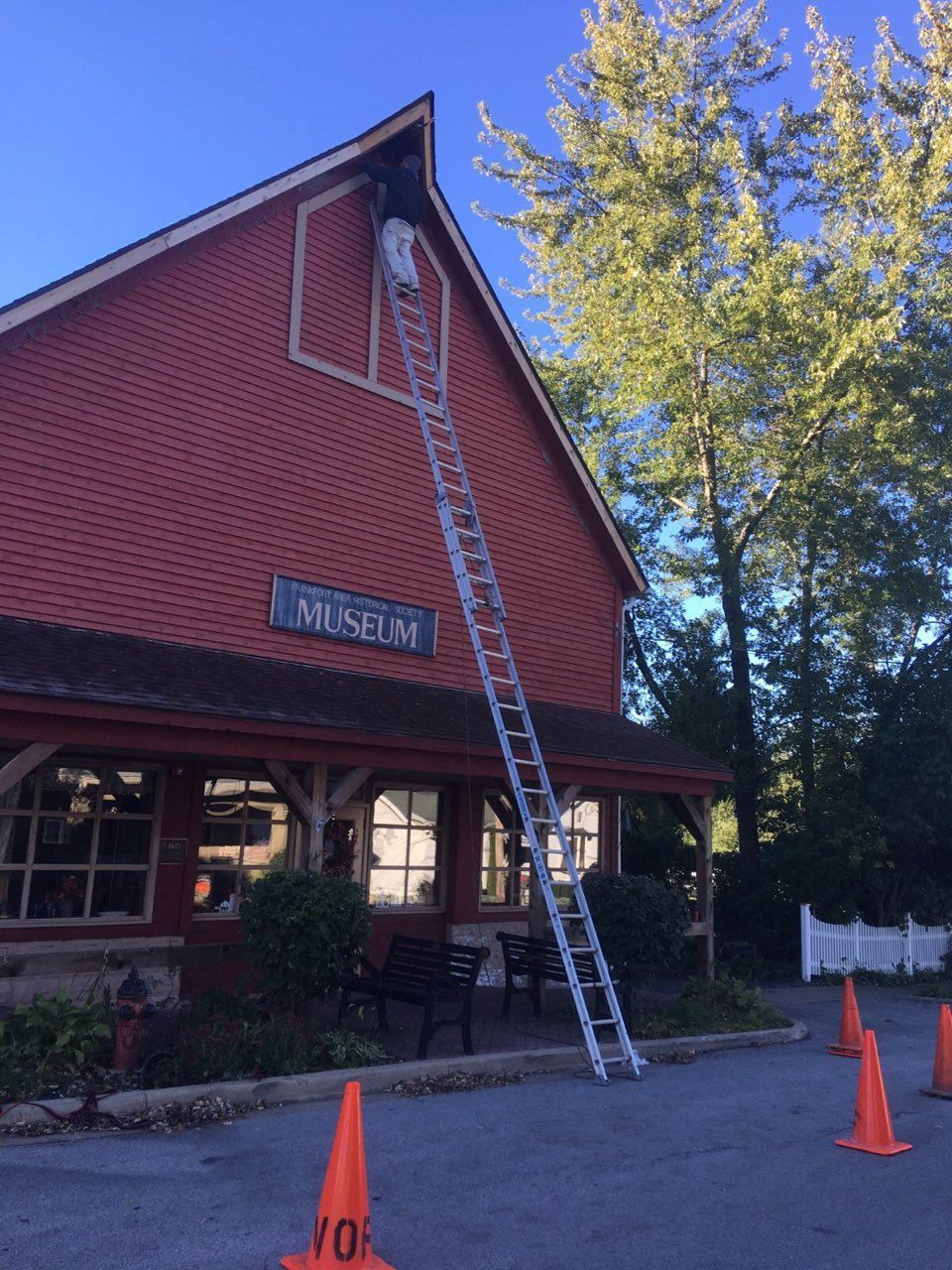 Ladder leaning to a red building roof, with orange cones and tall trees outside.
