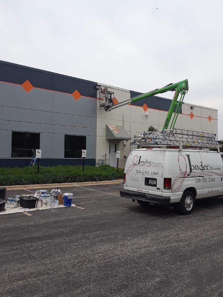 Worker in a green boom lift fixing the exterior of a gray industrial building beside a white van.