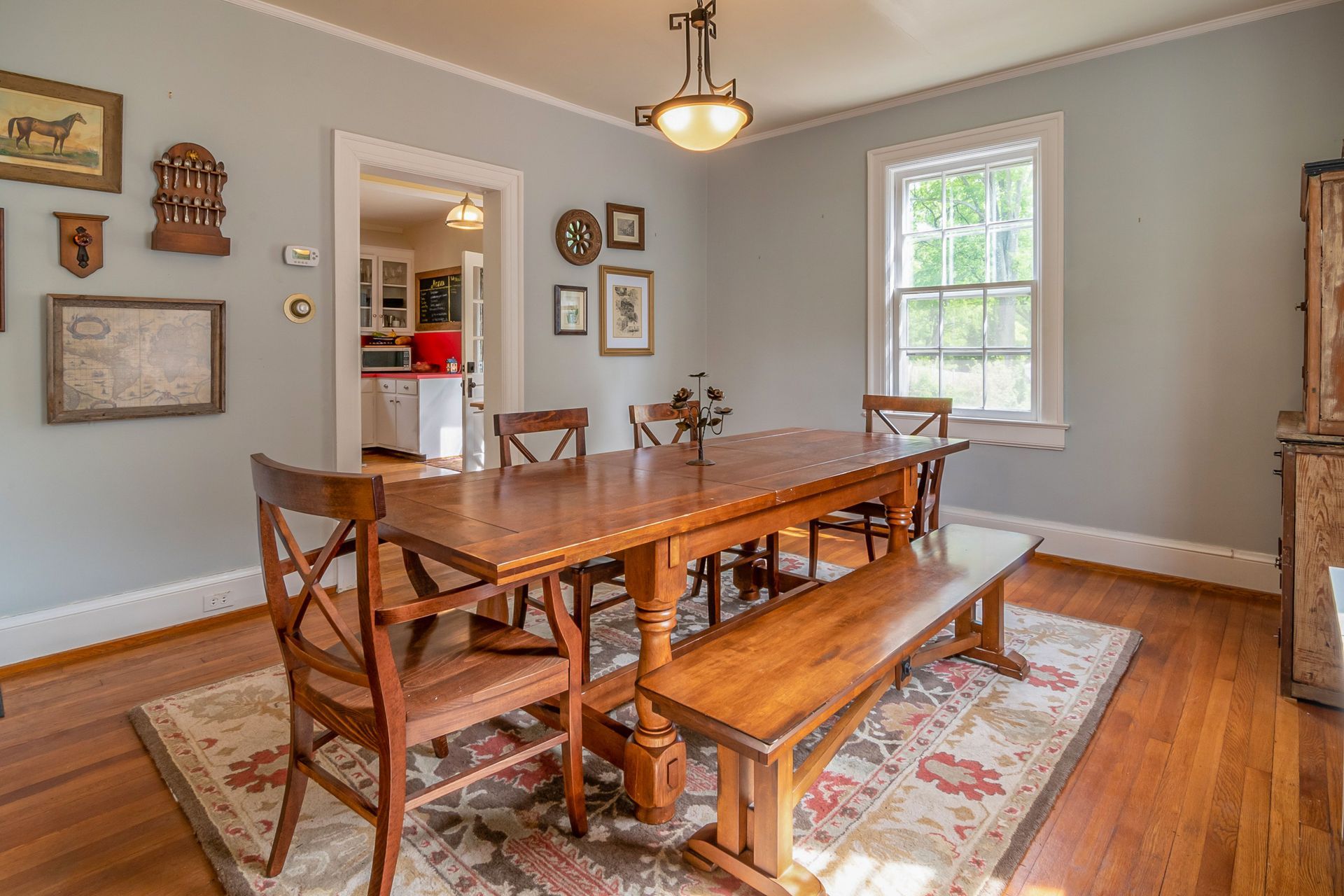 Dining room with long wooden table, bench, patterned rug, and light gray walls with framed decor.