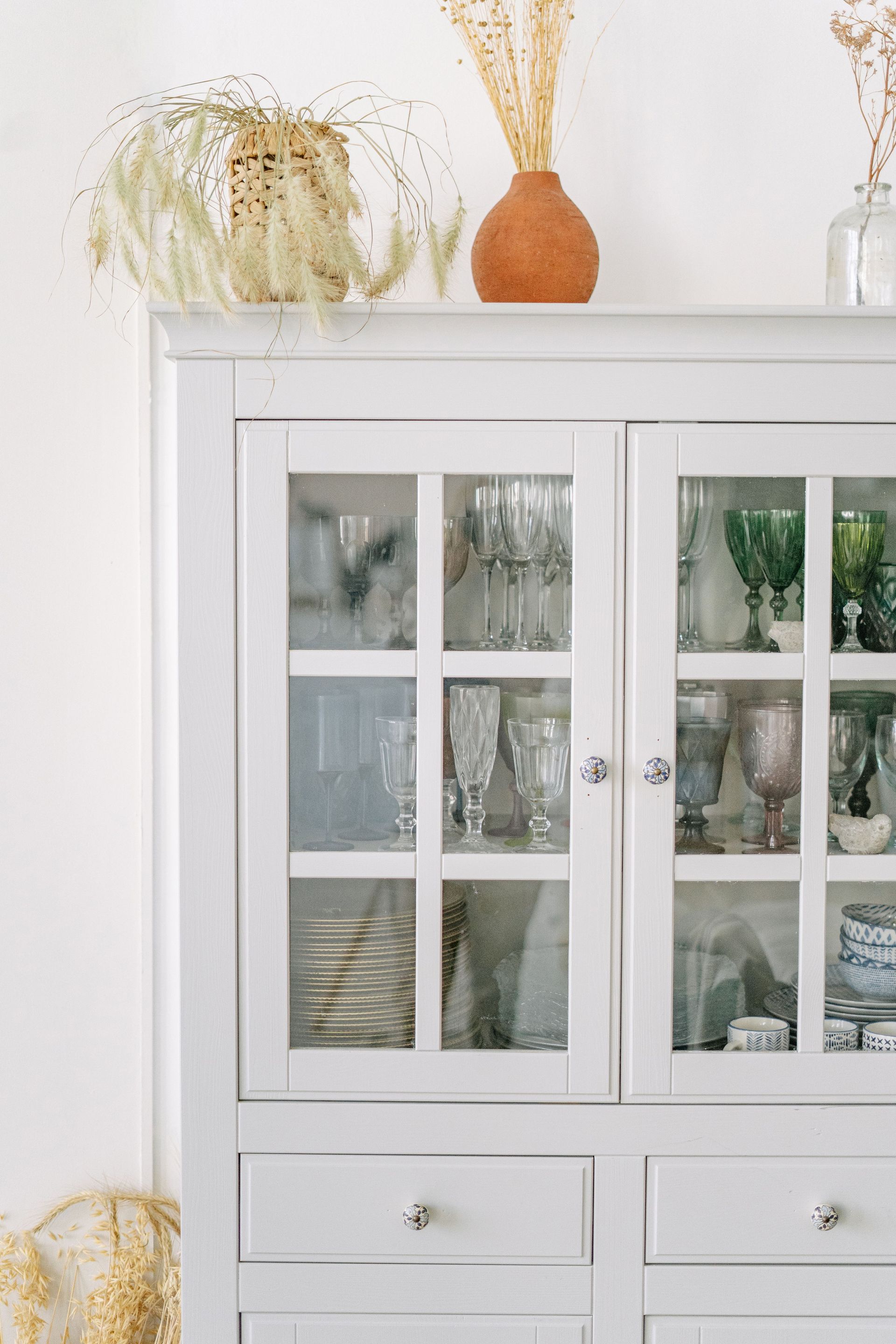 White glass-front cabinet with drawers, decorated with dried flowers and vases on top.