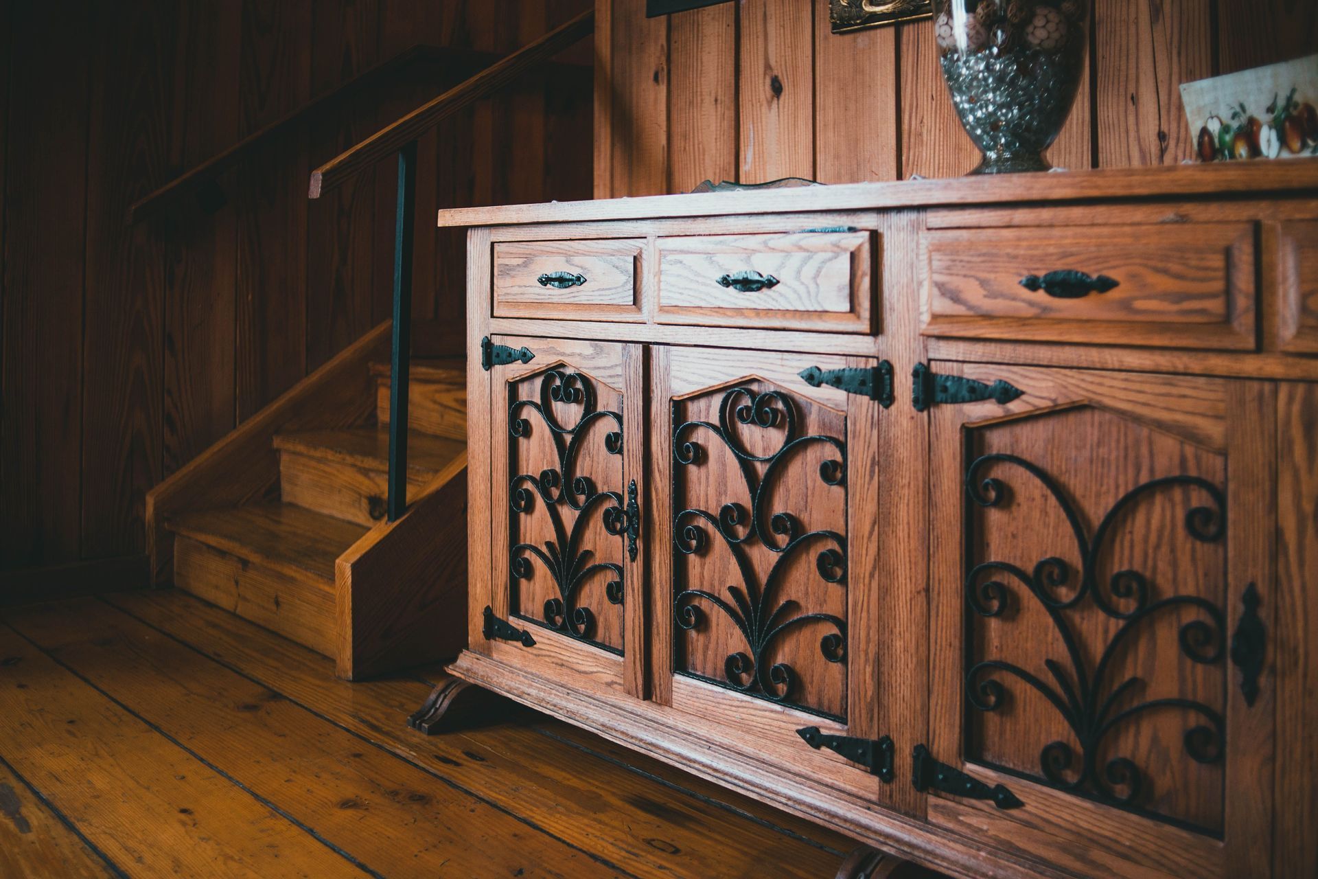 Wooden cabinet with wrought-iron doors beside stairs in a rustic wood-paneled room