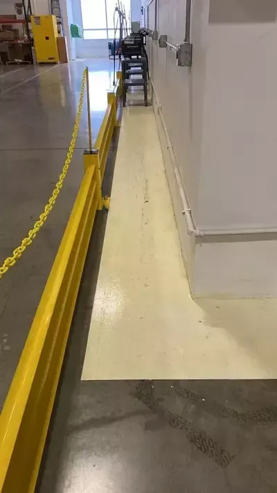 Narrow indoor hallway with a yellow guardrail beside a beige ramp and stacked chairs in the distance