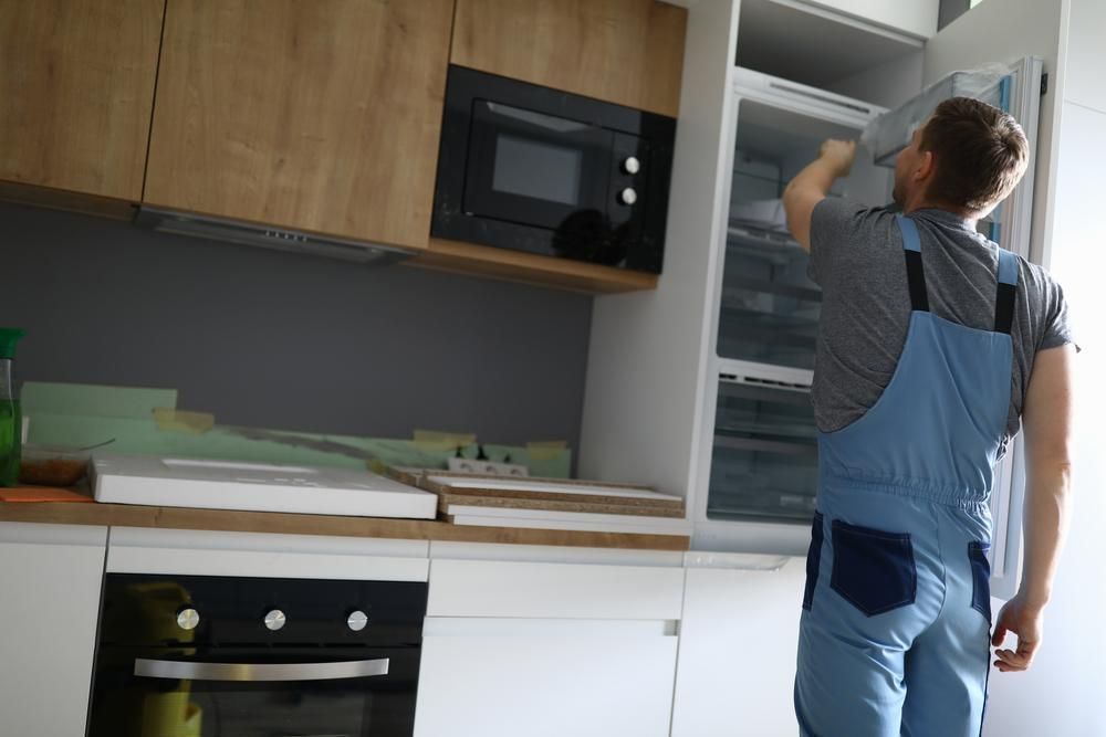 A Man In Blue Overalls Is Opening A Refrigerator Door In A Kitchen — Gold Coast City Plumbing In Helensvale, QLD