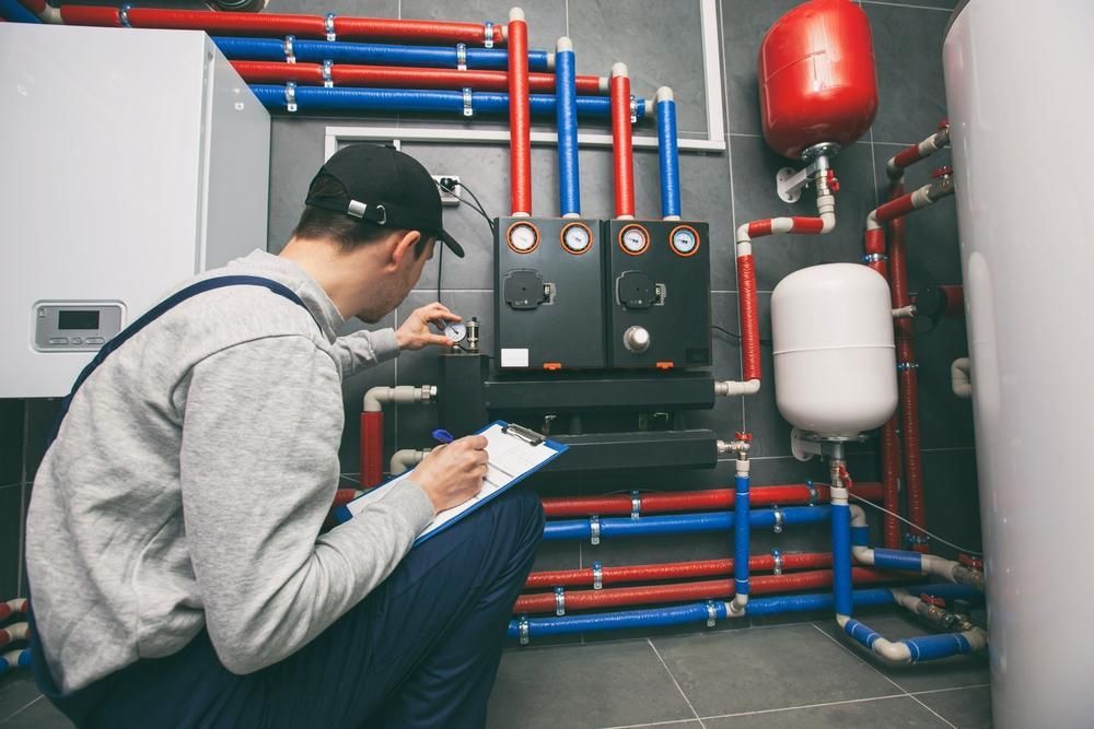 A Man Is Kneeling Down In A Room With Pipes And Writing On A Clipboard — Gold Coast City Plumbing In Robina, QLD