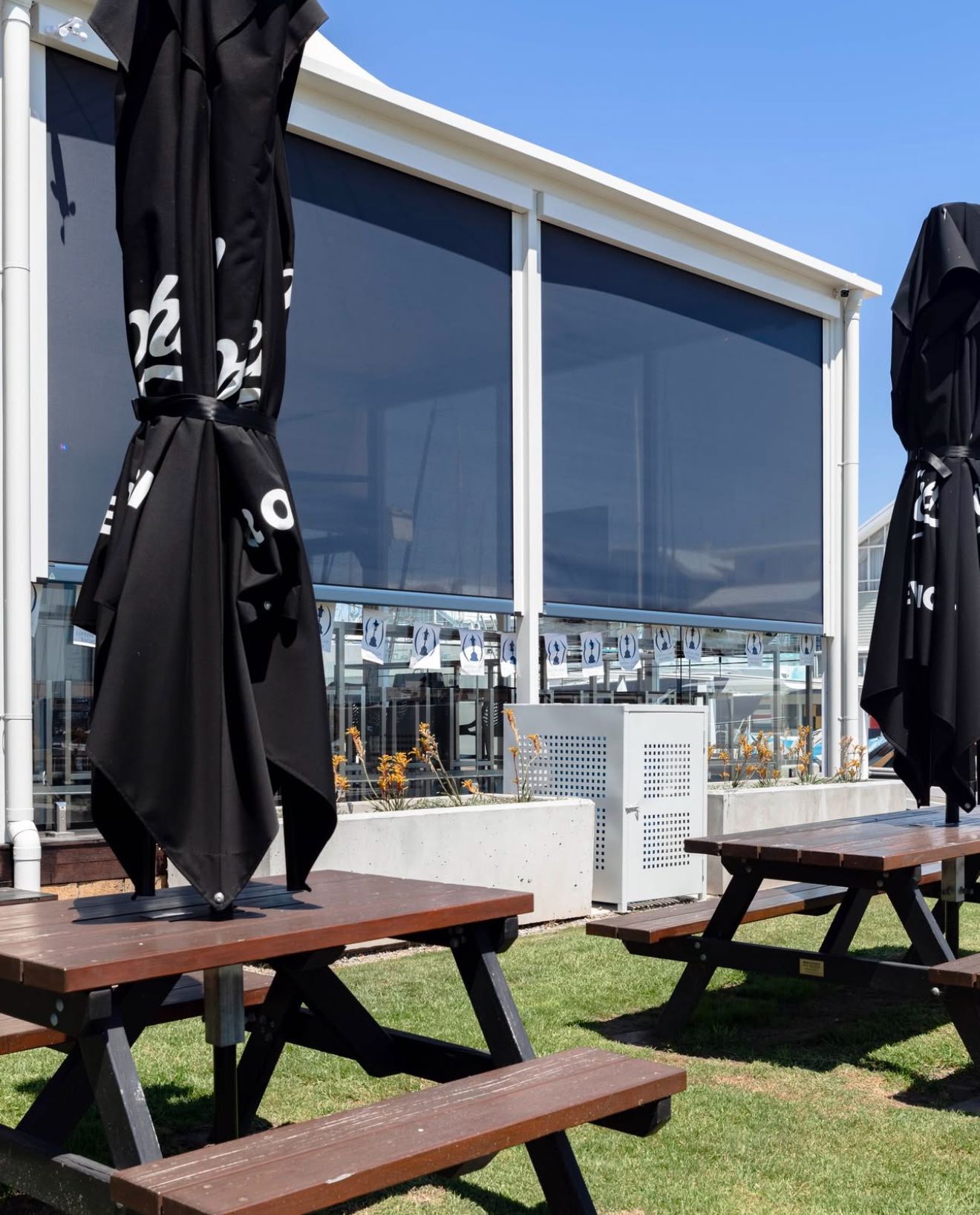 Outdoor restaurant with picnic tables, closed black umbrellas, and shaded windows.