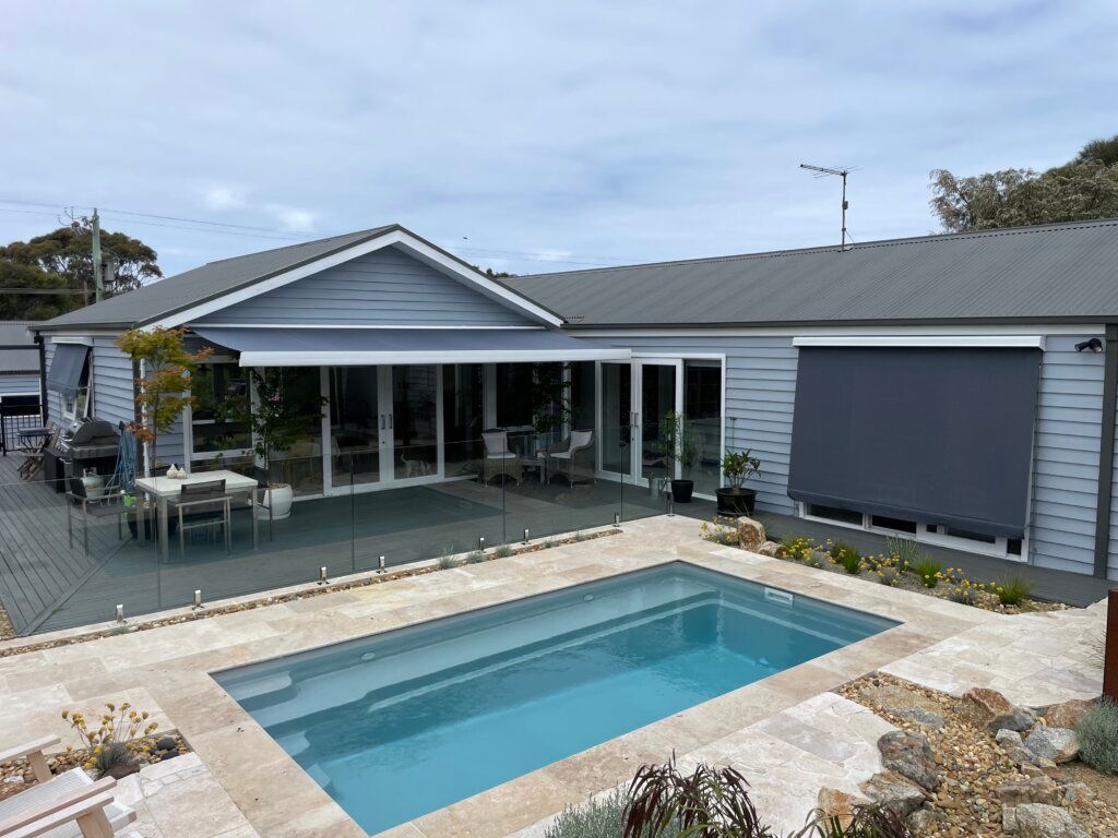 A home with a pool, patio, and glass fence. Blue siding, gray roof, and retractable shades are visible.