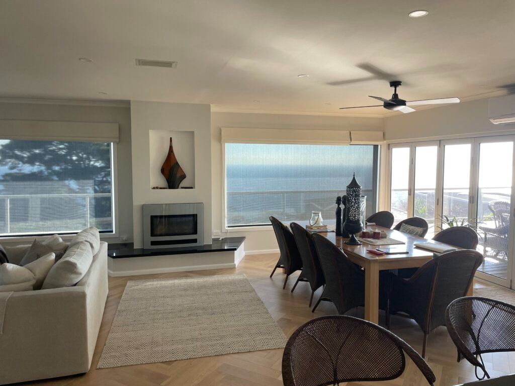 Living room with ocean view, fireplace, dining table, beige sofa, rug, wood flooring, and large windows.