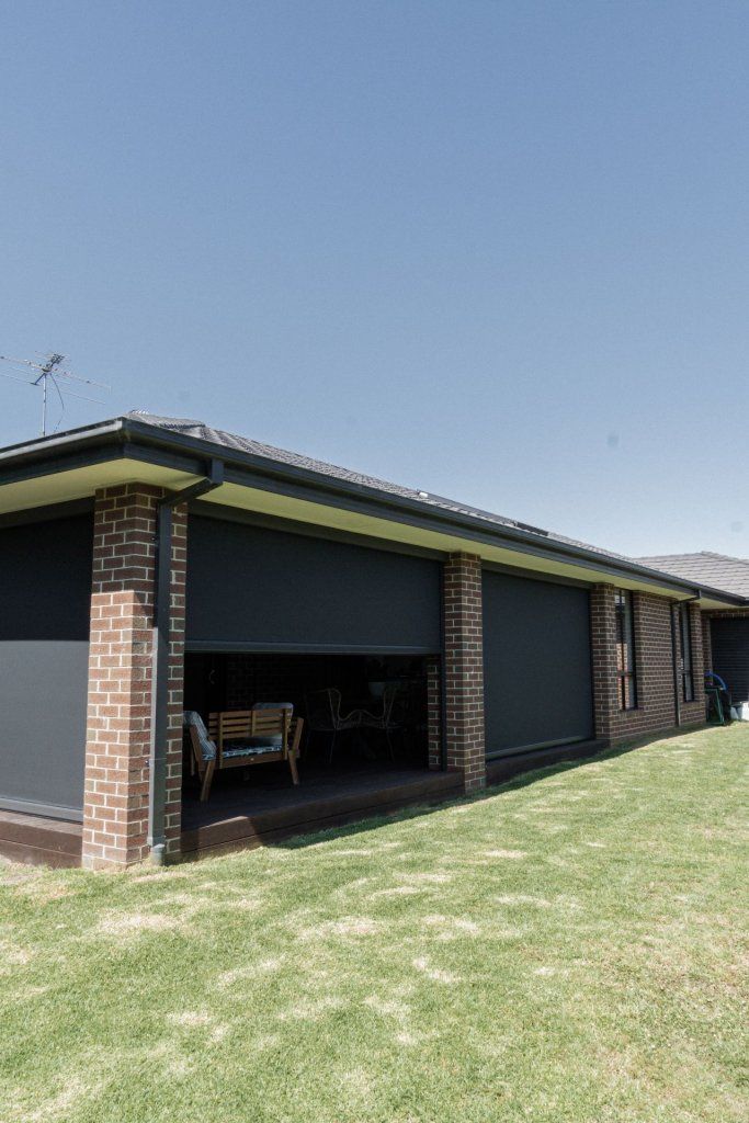 Dark gray outdoor blinds on a brick patio, attached to a house with a green lawn and blue sky.