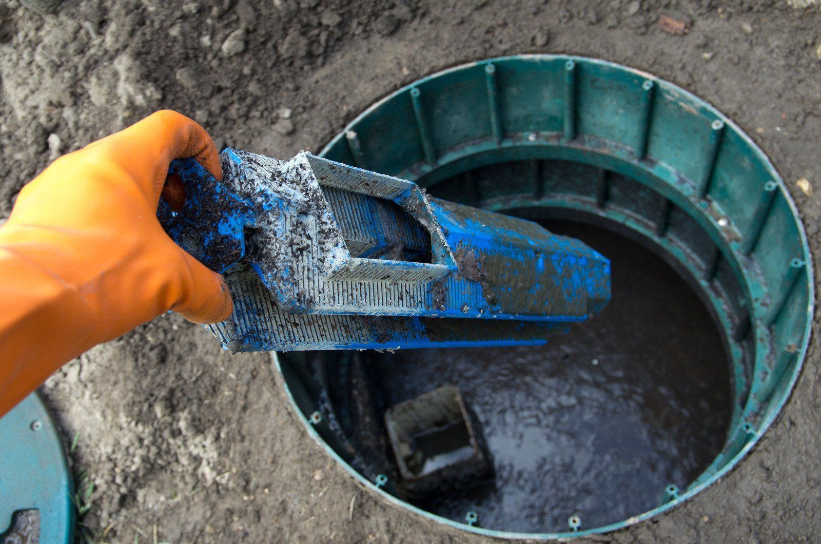Gloved hand lowering a blue filter into a green manhole opening with standing water