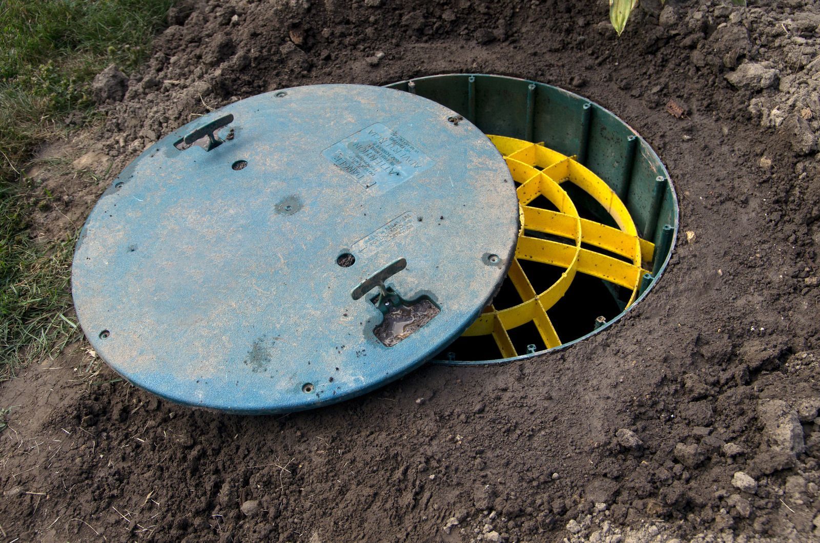 Blue septic tank lid open, revealing yellow interior structure, in a yard.
