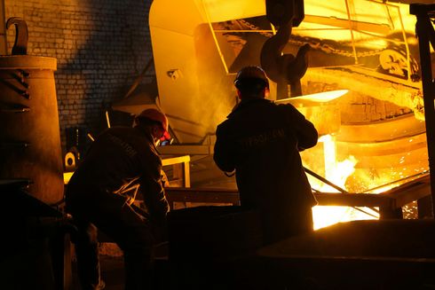 Two workers in protective gear at a steel mill, pouring molten metal. Bright orange glow, dim setting.