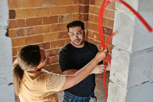 Two people installing red electrical conduit in a building under construction, near brick walls, wearing safety glasses.