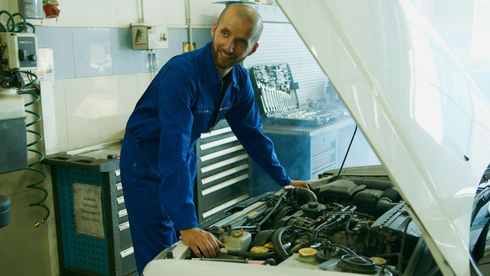 Mechanic in blue overalls working on a white car engine, smiling in a garage.