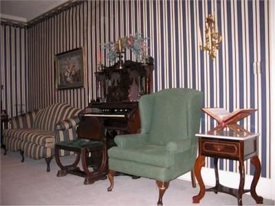 Living room with striped wallpaper, featuring a sofa, chair, antique organ, and side table.