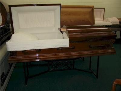 Open, wooden casket with white interior displayed on a table. Additional caskets in background.