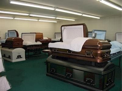 Interior of a funeral home displaying several open and closed caskets on a green floor under bright lights.