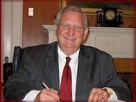 Man in suit signing document at a desk with a clock and fireplace in the background.