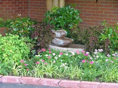 Fountain surrounded by plants and flowers in front of a brick building.