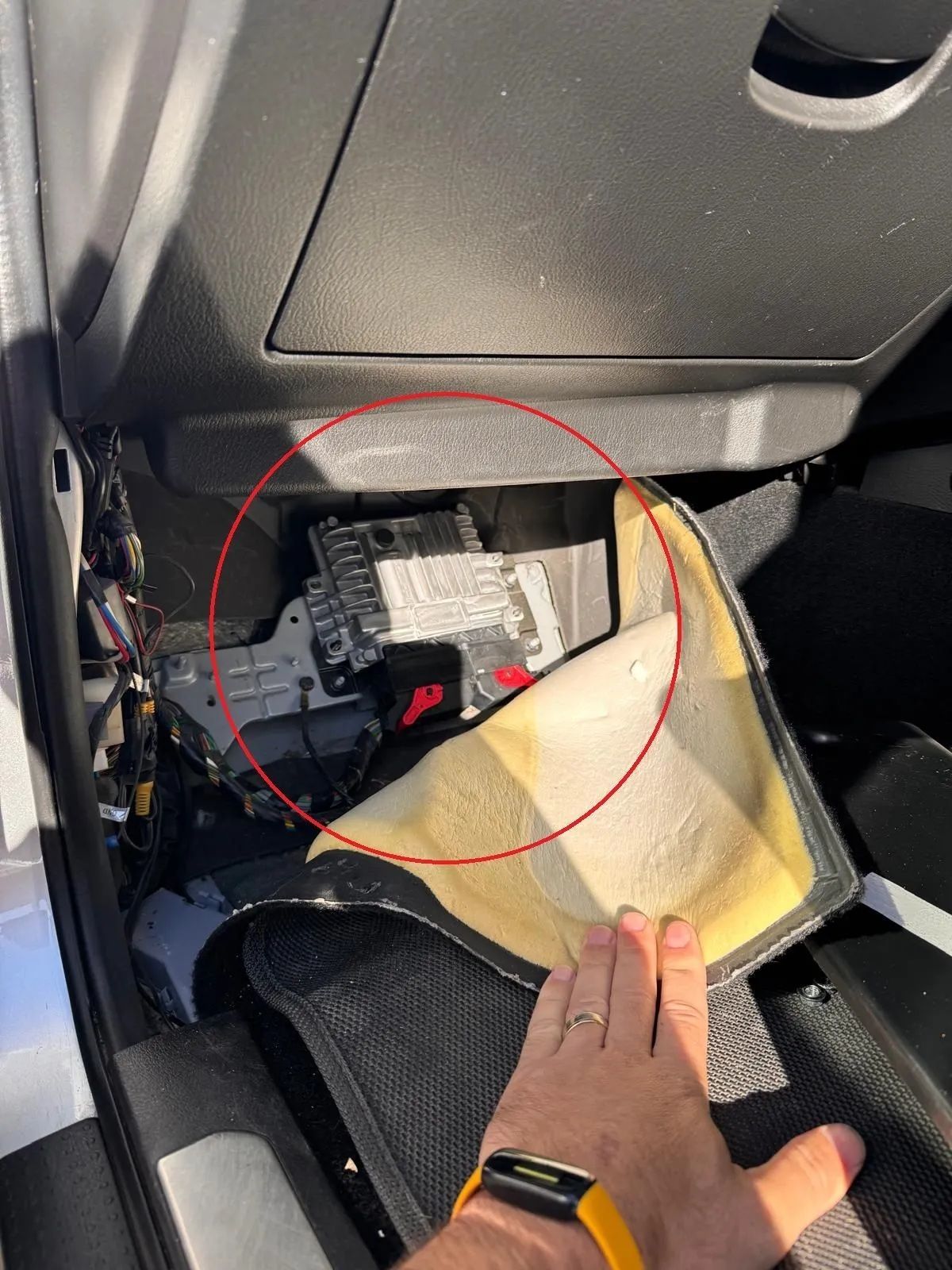 Hand pointing at a red-circled car interior panel with exposed wiring and a loose beige mat.