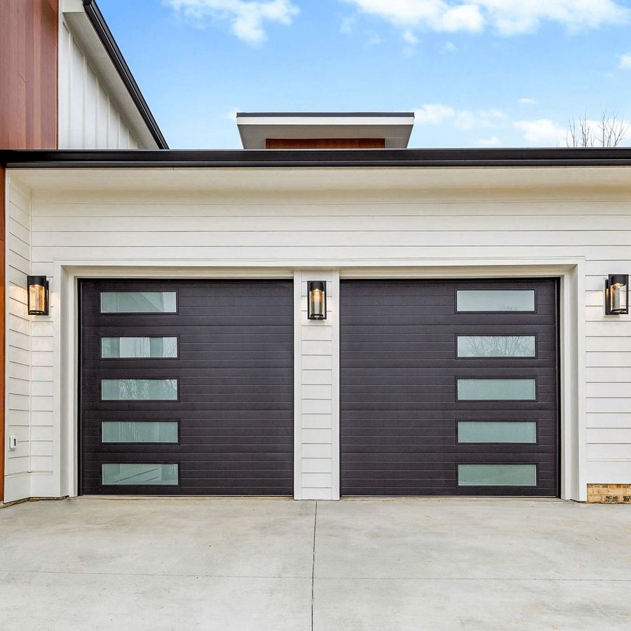 Two dark gray garage doors with rectangular glass panels, white siding, and outdoor lights.