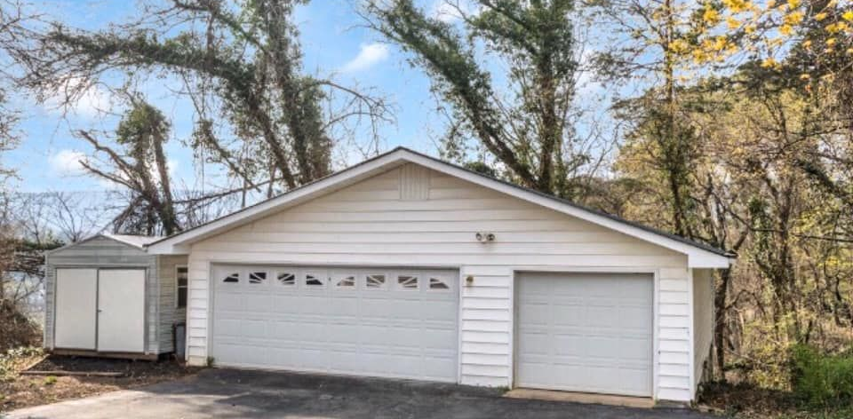White garage with two doors and a small shed, surrounded by trees and a cloudy sky.
