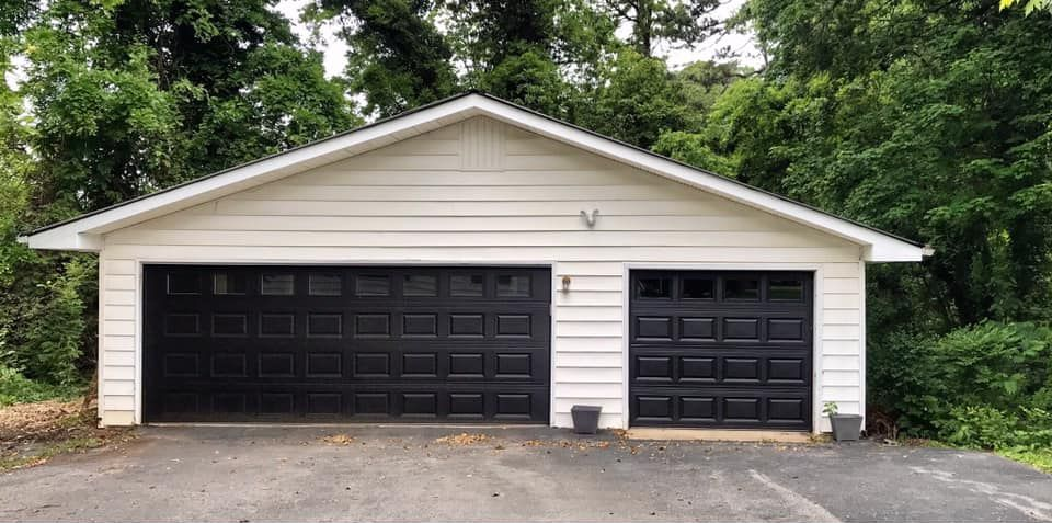White garage with two black garage doors, against a backdrop of trees.