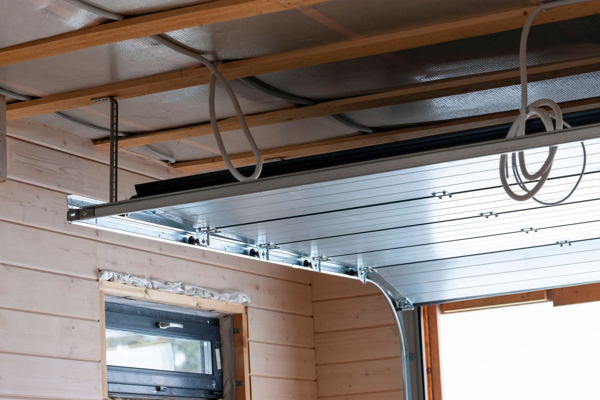 Garage door installation, showing metal door panel, wooden beams, wiring, and support brackets.