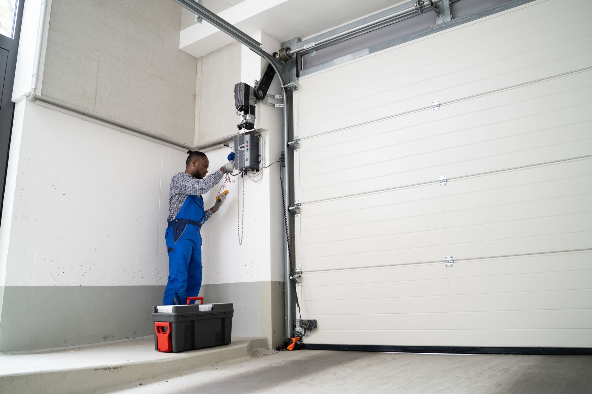 Man in blue overalls repairs garage door opener, standing on a toolbox inside a garage.