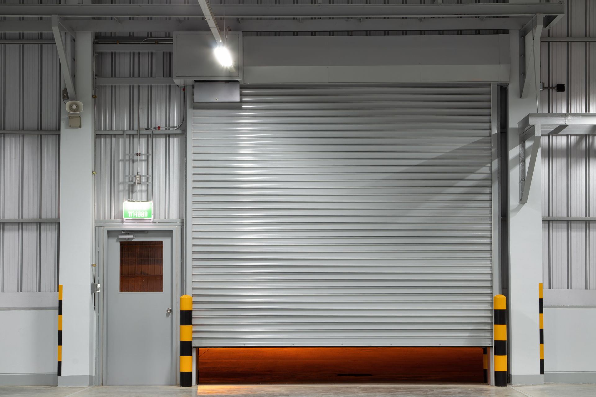 Rolling gray warehouse door with a closed metal shutter, beside a door and safety poles.