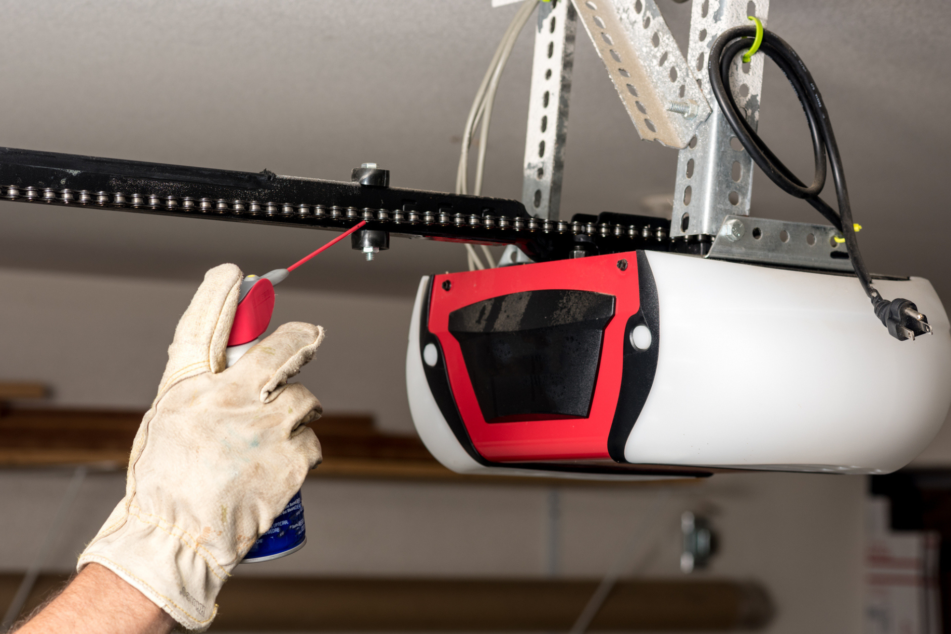 Person wearing gloves, spraying lubricant on a garage door opener chain.