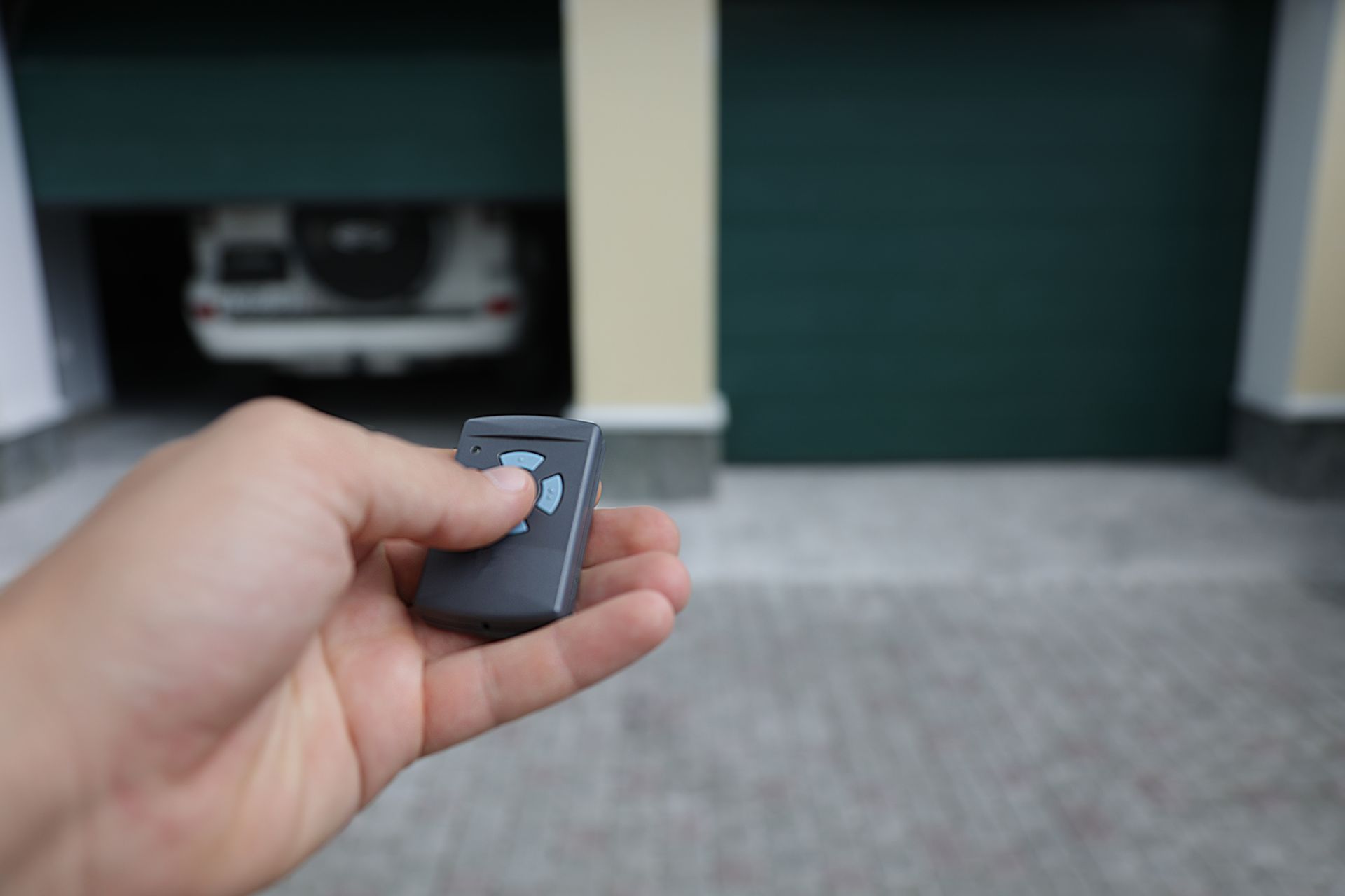 Hand holding a garage door opener, opening a dark green garage door with a car visible inside.