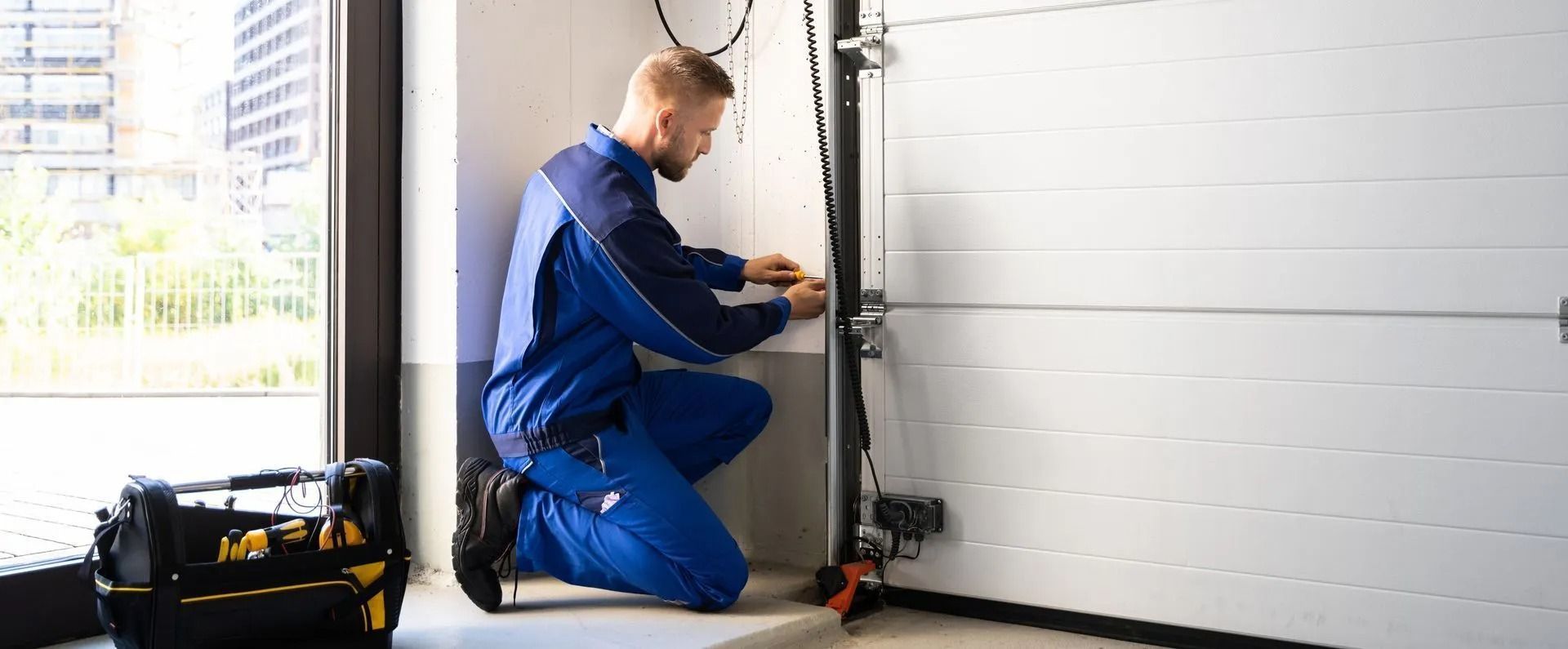 Technician in blue overalls kneeling to adjust a garage door opener beside a white sectional door.