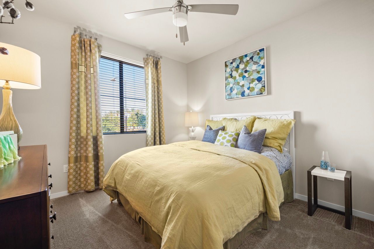 Bedroom in an apartment with yellow bedding, a dresser, nightstand, and window with patterned curtains.