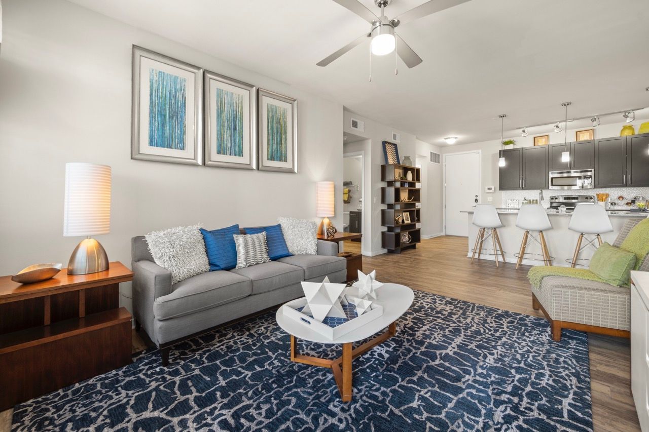 Open-concept living room with gray sofa, blue pillows, and a modern kitchen with white stools.