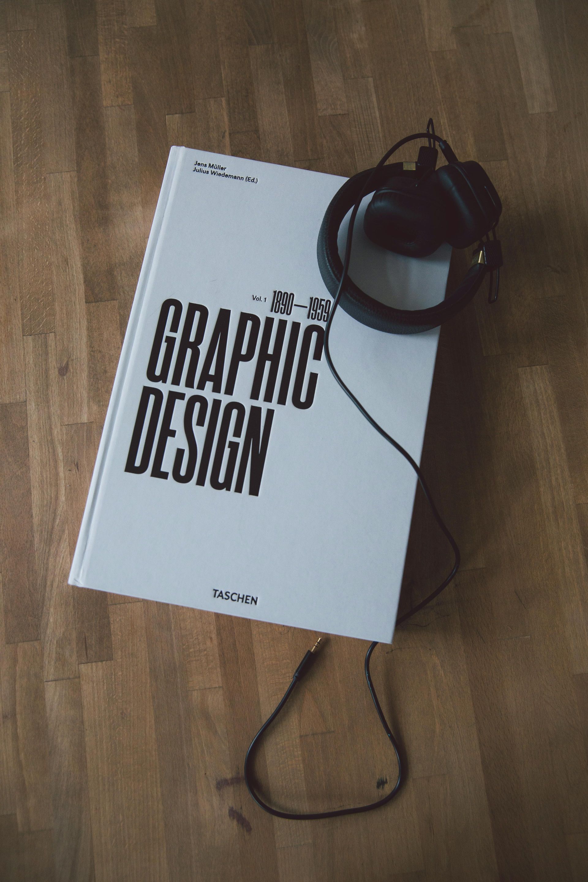 A book about graphic design is sitting on a wooden floor next to a pair of headphones.