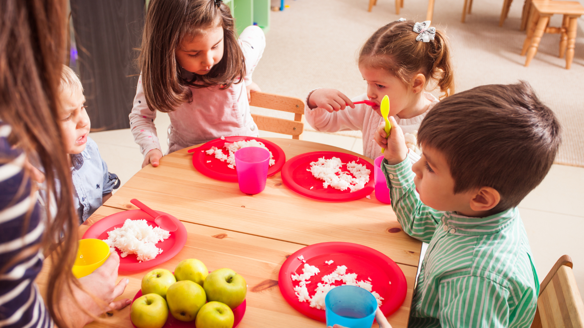 Two teachers hugging several young children in a brightly lit classroom.