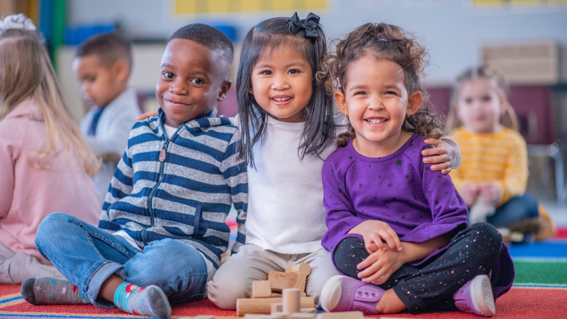 Three diverse children smile, sitting closely together on a rug in a classroom.