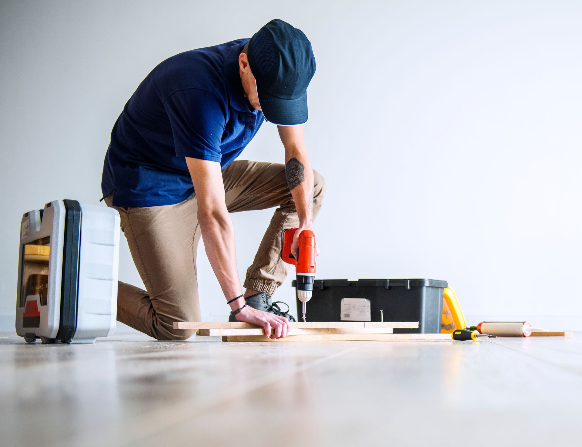 A man is kneeling on the floor using a drill on a piece of wood.