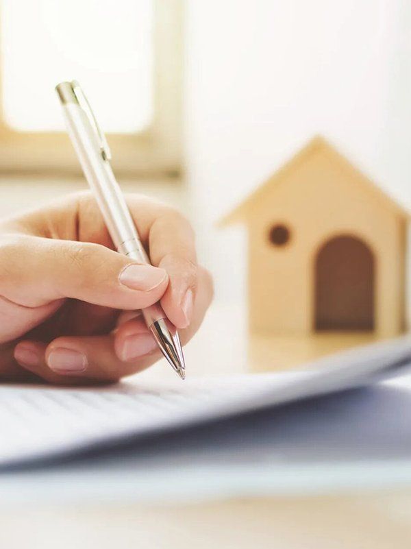 Woman Holding A Pen With Mini House In The Background — Outback Conveyancing Services in Alice Springs, NT