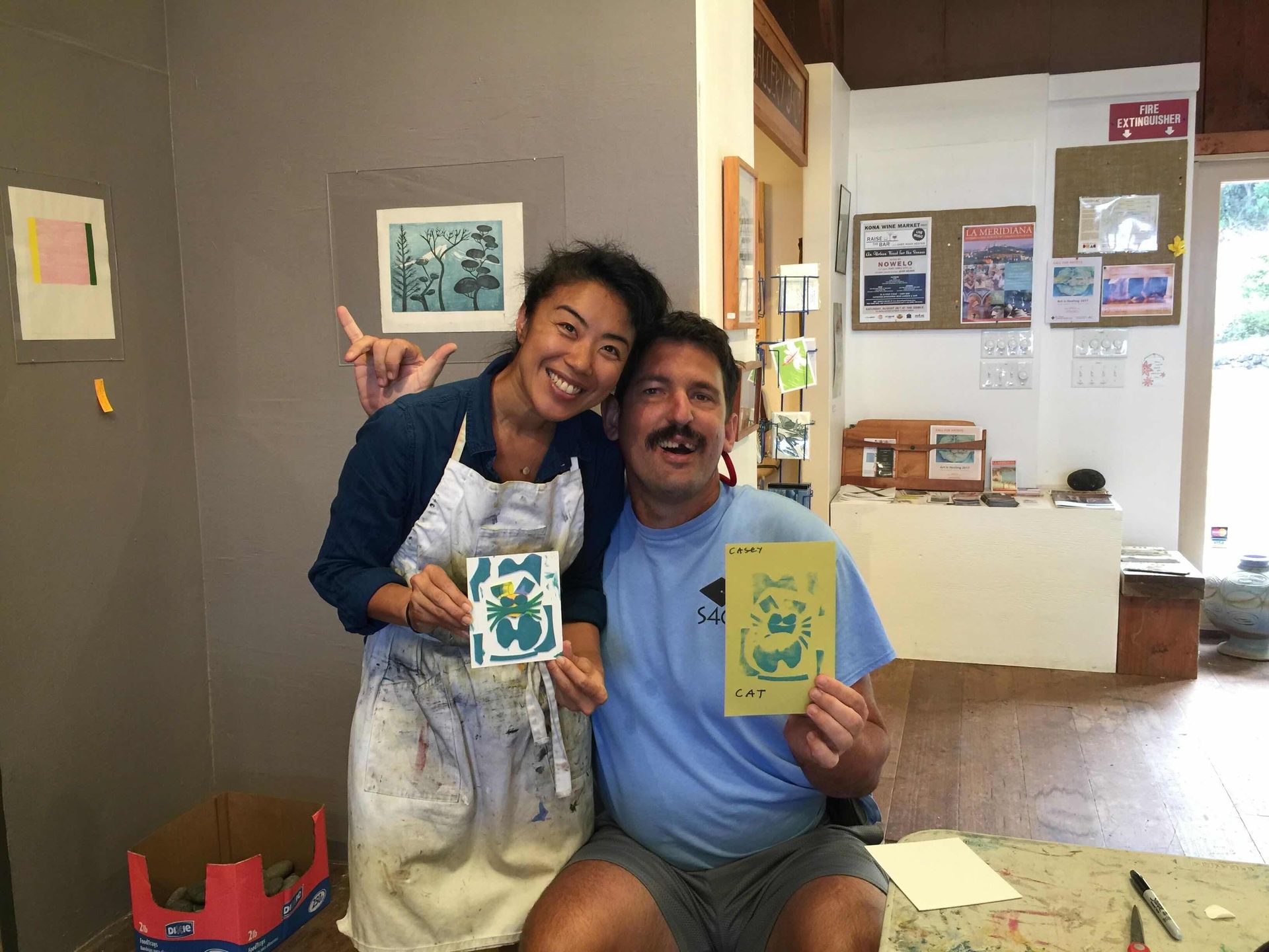 A smiling duo in an art studio, each holding a small, decorated card. The person on the left wears a paint-splattered apron.