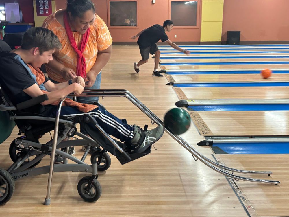 A woman is helping a boy in a wheelchair bowl at a bowling alley.