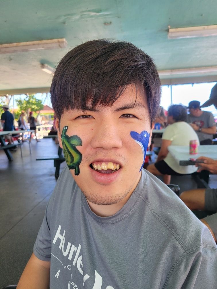 A man with a cactus painted on his face is sitting at a picnic table.