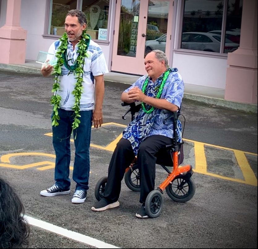 A man wearing a lei is standing next to a man in a wheelchair