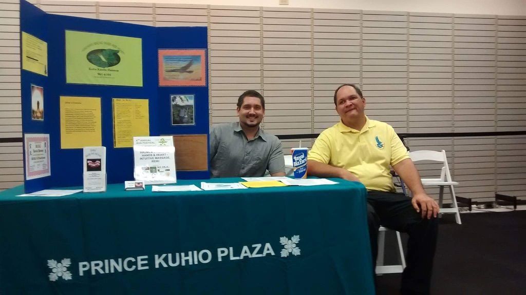 Two men sit at a table with a blue table cloth that says prince kunio plaza