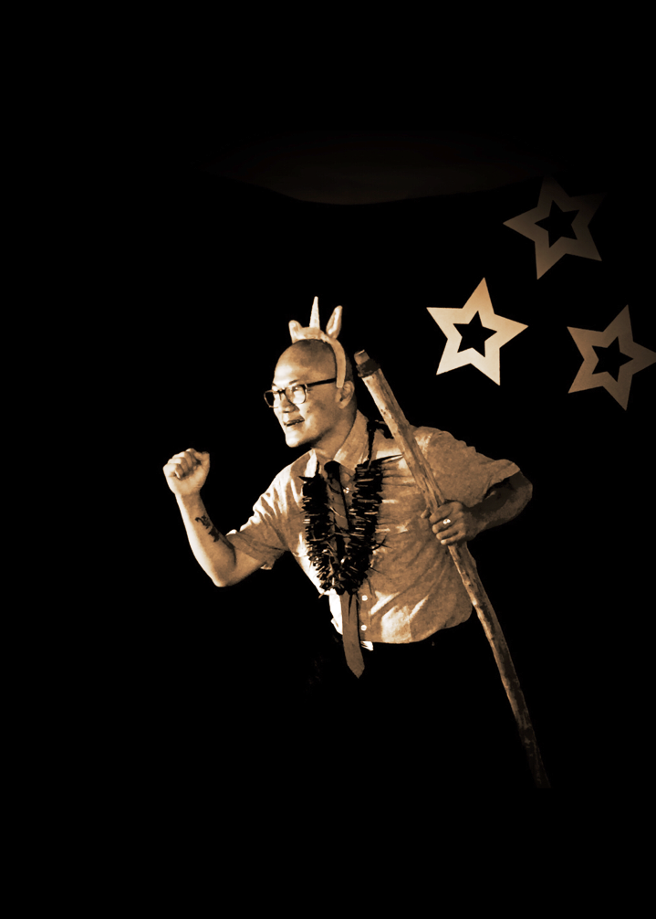 A man wearing glasses and a lei is holding a stick in the dark.