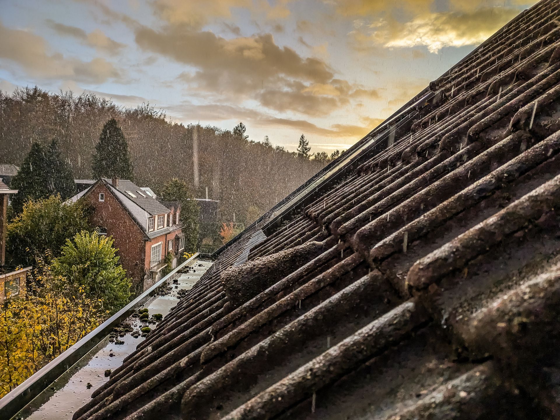 Rain falling on a tiled roof, overlooking a town with trees and buildings, under a cloudy sky.