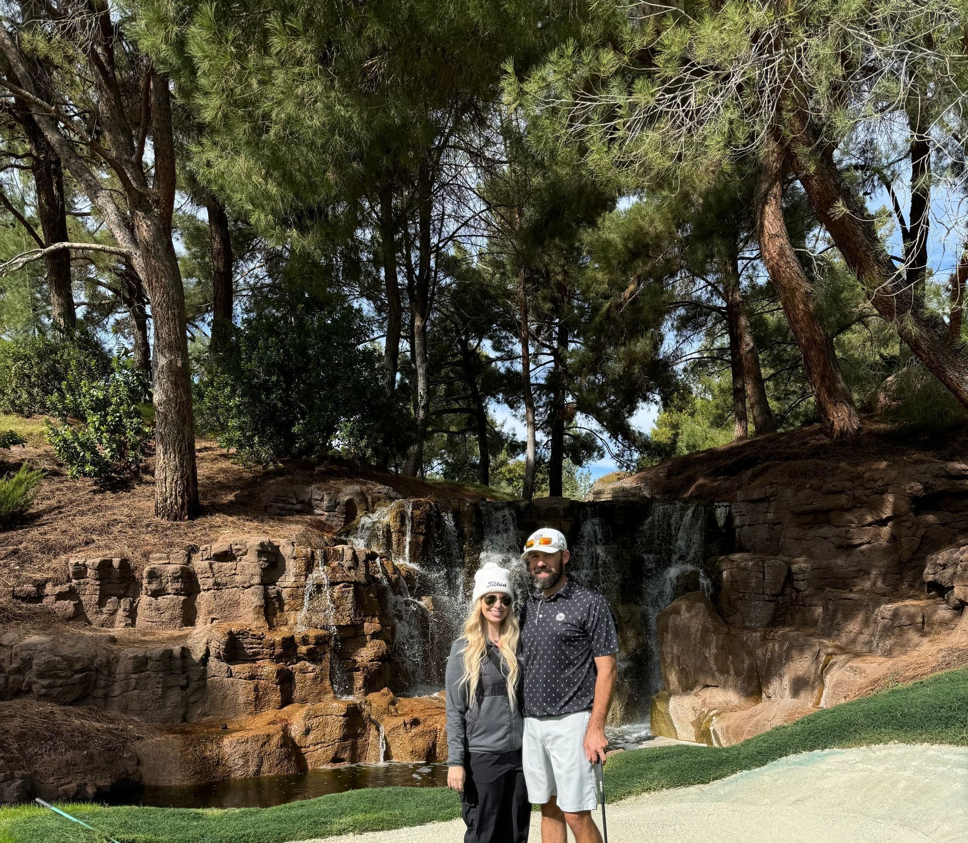 A man and a woman are standing on a golf course in front of a waterfall.