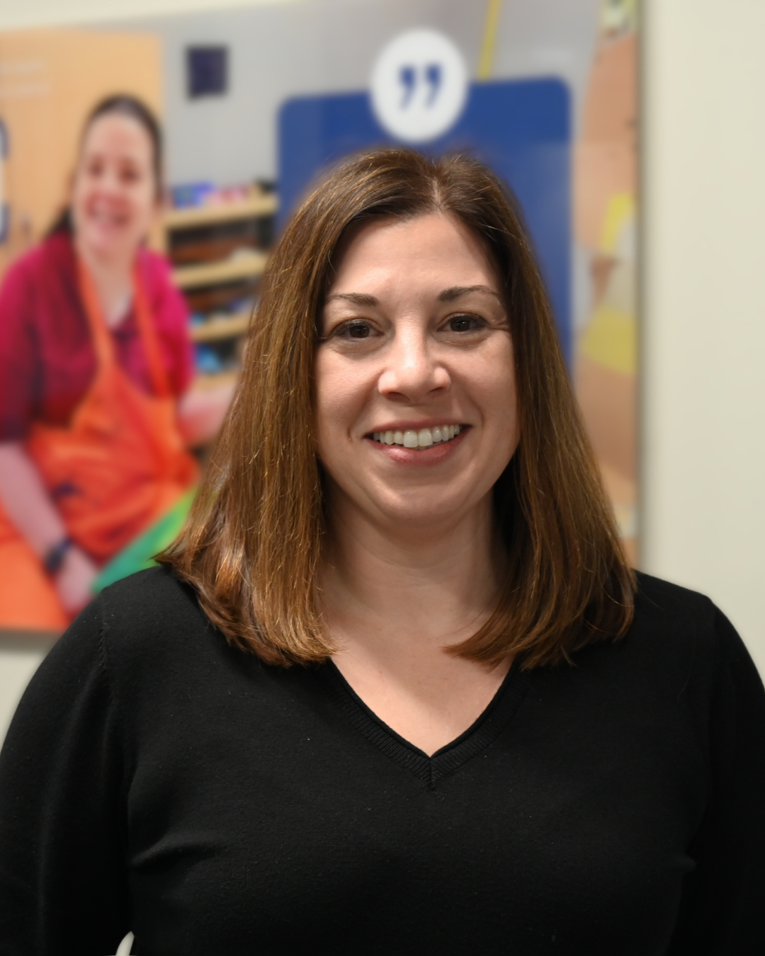 Woman with brown hair smiles, wearing a black top against a blurred gray background.